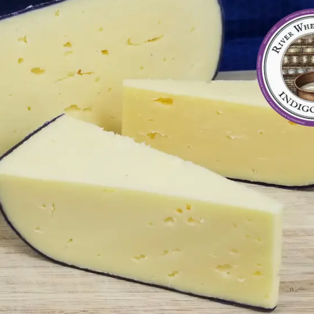 Wedges of Indigo Ridge artisan cheese from River Whey Creamery displayed on a wooden surface, showing a pale yellow interior with small holes and a natural dark rind, presented as a gourmet handcrafted cheese.