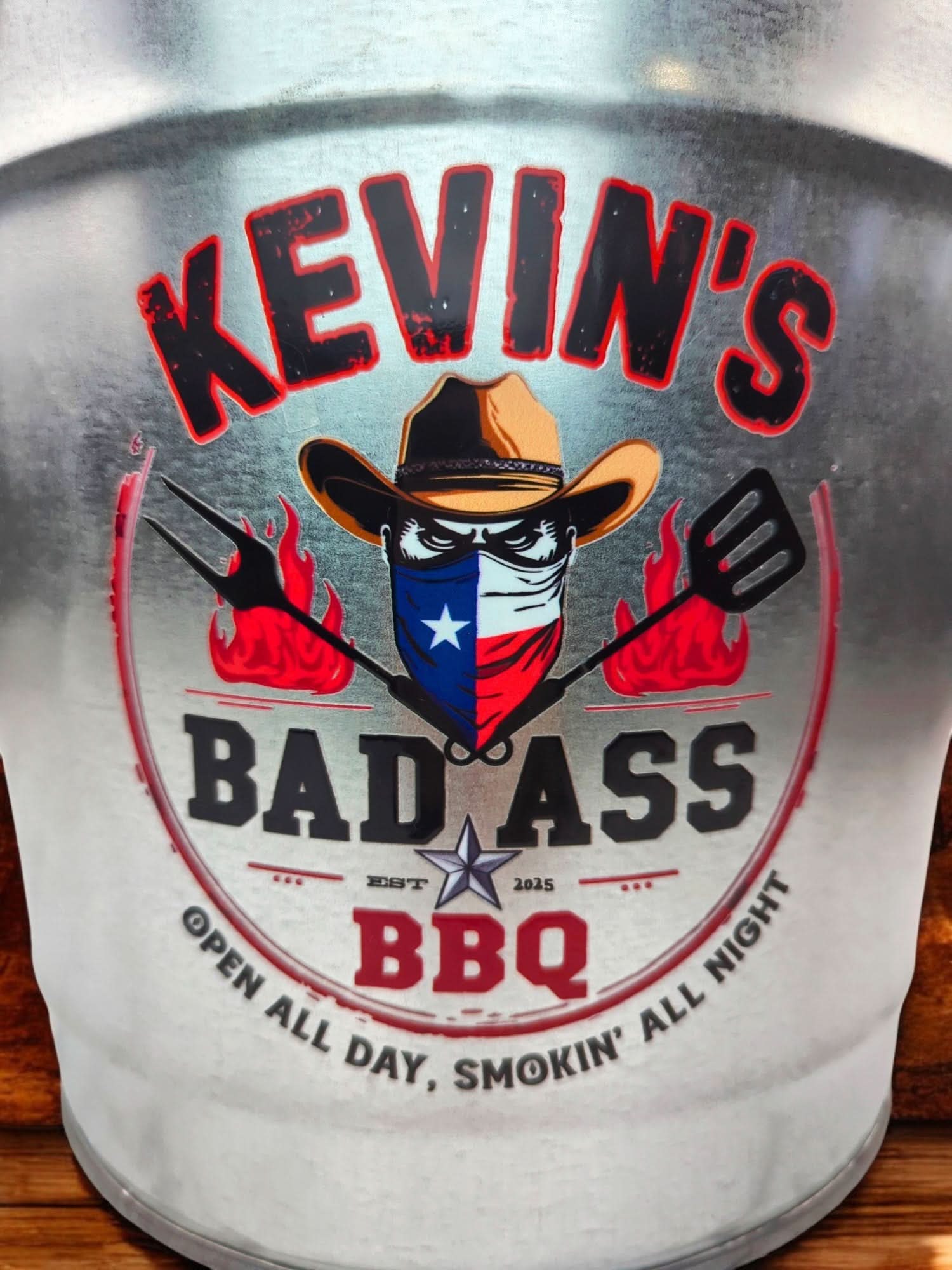 Close-up of a custom “Kevin’s Bad Ass BBQ” logo printed on drinkware, featuring a cowboy-hat character with a Texas bandana, crossed grill tools, flames, and bold barbecue branding text.