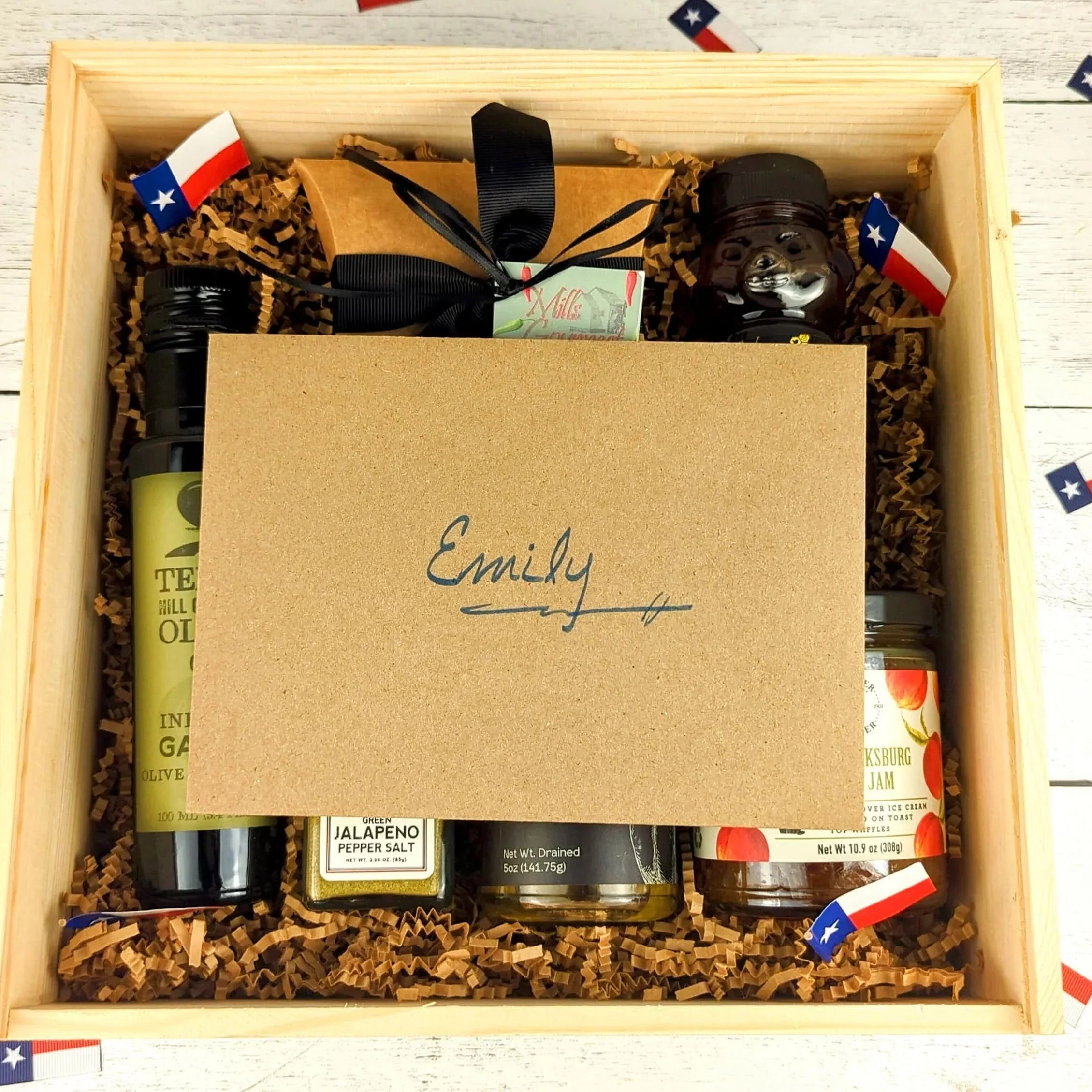 Open wooden gift box filled with Texas-made gourmet items, including infused olive oil, jam, seasoning, and sweets, topped with a personalized kraft card addressed to Emily, arranged with shredded paper filler and Texas flag confetti.