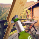 Bamboo grill scraper with a built-in metal bottle opener removing the cap from a green glass bottle on a rustic outdoor cabin deck, with wooden furniture and a mountain landscape in the background.