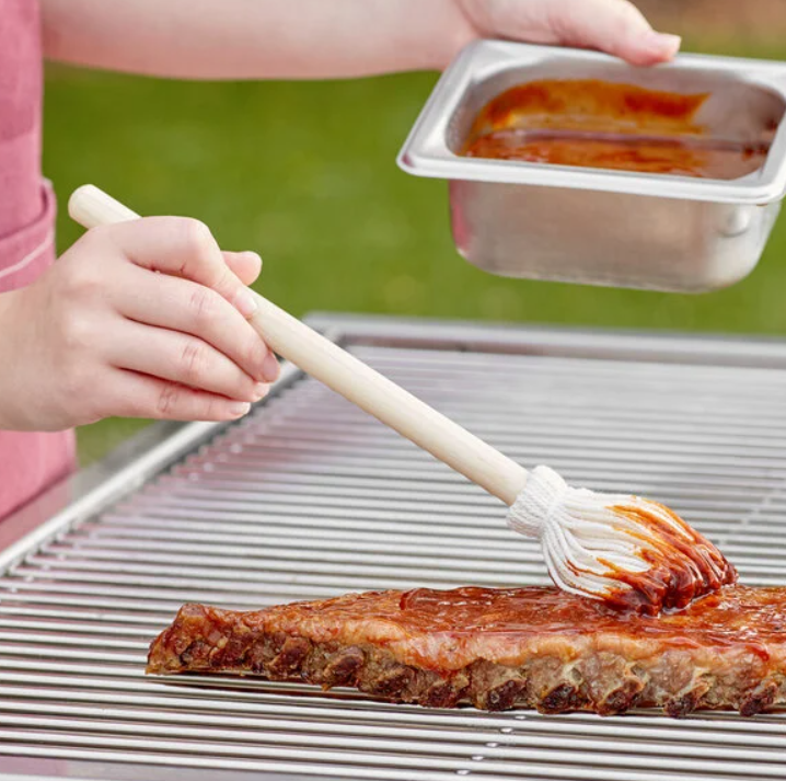 Close-up of barbecue ribs being brushed with sauce on a metal grill, with a hand holding a basting brush and a small container of barbecue sauce during outdoor cooking.