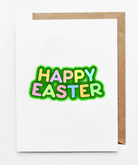 Happy Easter greeting card featuring playful pastel lettering set in bright green grass on a white background with a kraft paper envelope