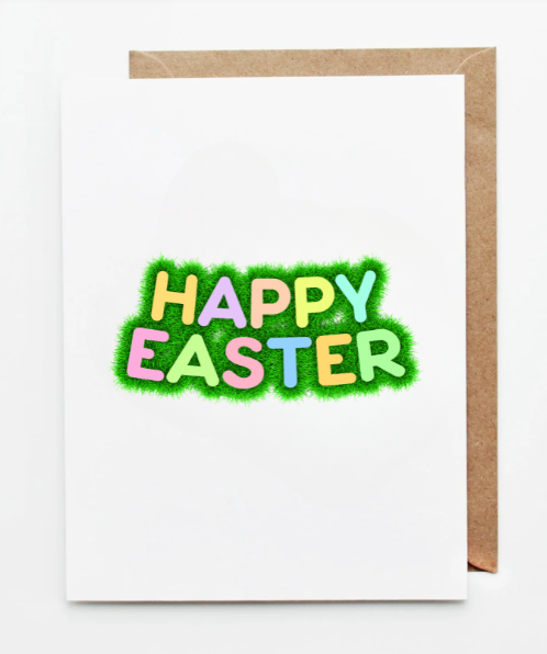 Happy Easter greeting card featuring playful pastel lettering set in bright green grass on a white background with a kraft paper envelope