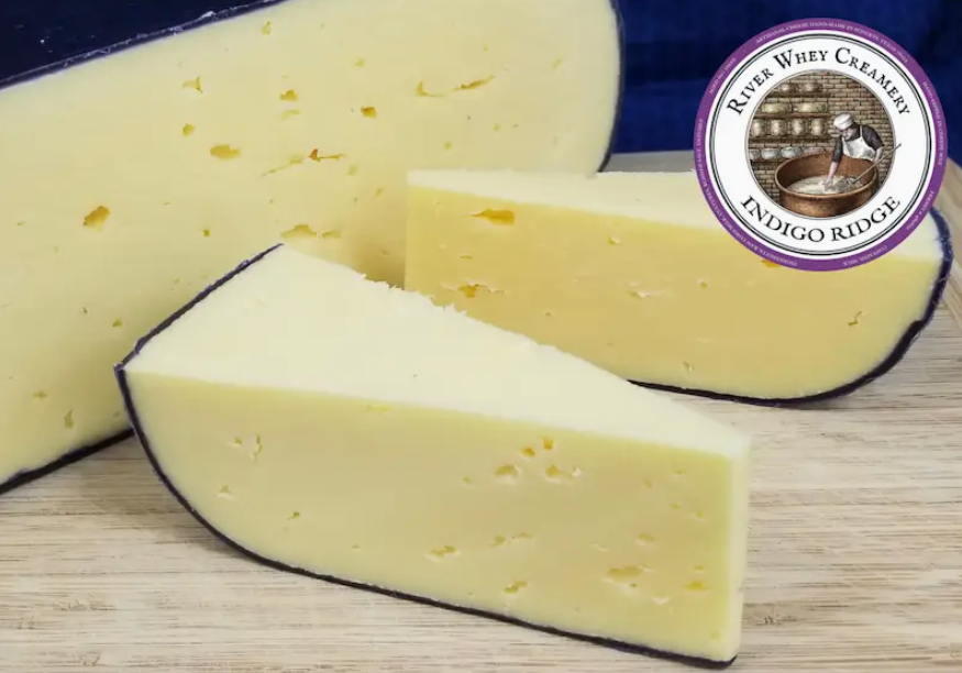 Wedges of Indigo Ridge artisan cheese from River Whey Creamery displayed on a wooden surface, showing a pale yellow interior with small holes and a natural dark rind, presented as a gourmet handcrafted cheese.