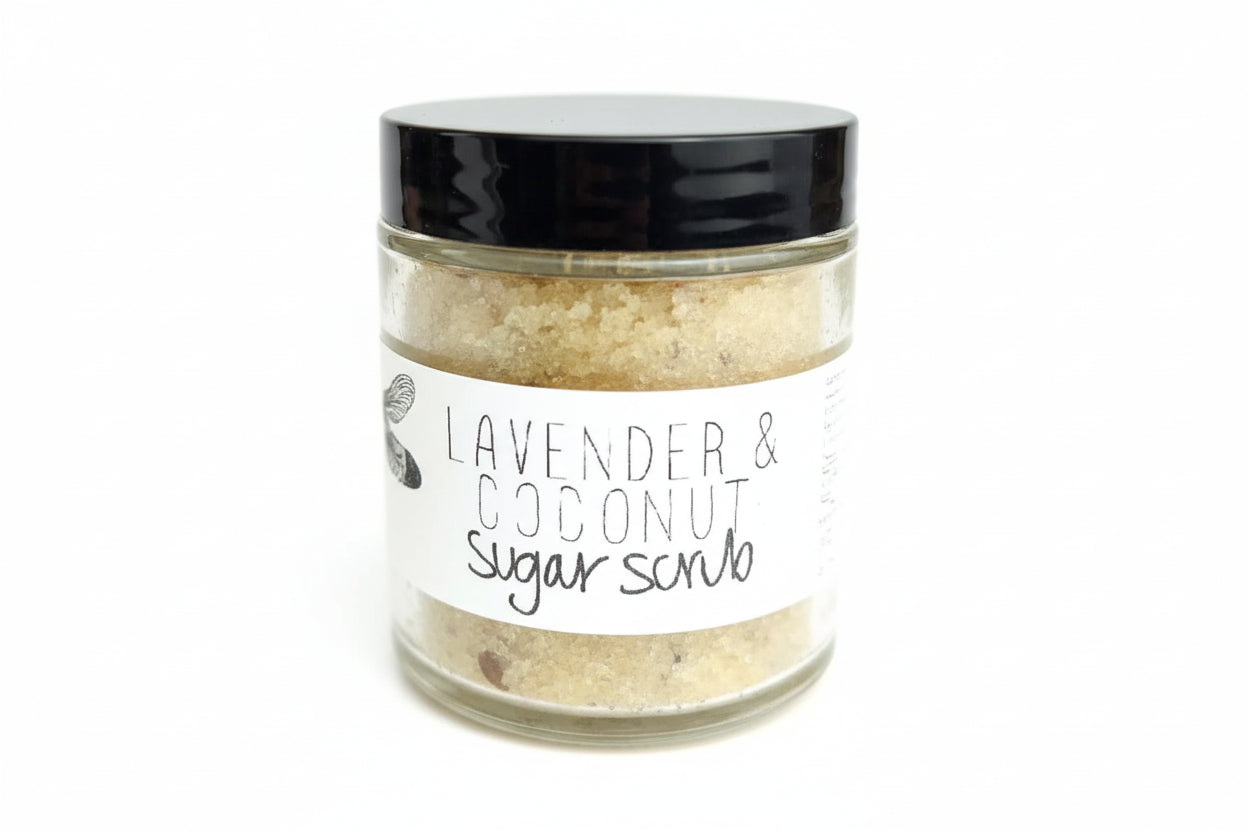 Glass jar of lavender and coconut sugar scrub with a black lid, showing textured sugar crystals and a minimalist label, displayed on a light rustic surface.