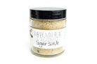 Glass jar of lavender and coconut sugar scrub with a black lid, showing textured sugar crystals and a minimalist label, displayed on a light rustic surface.