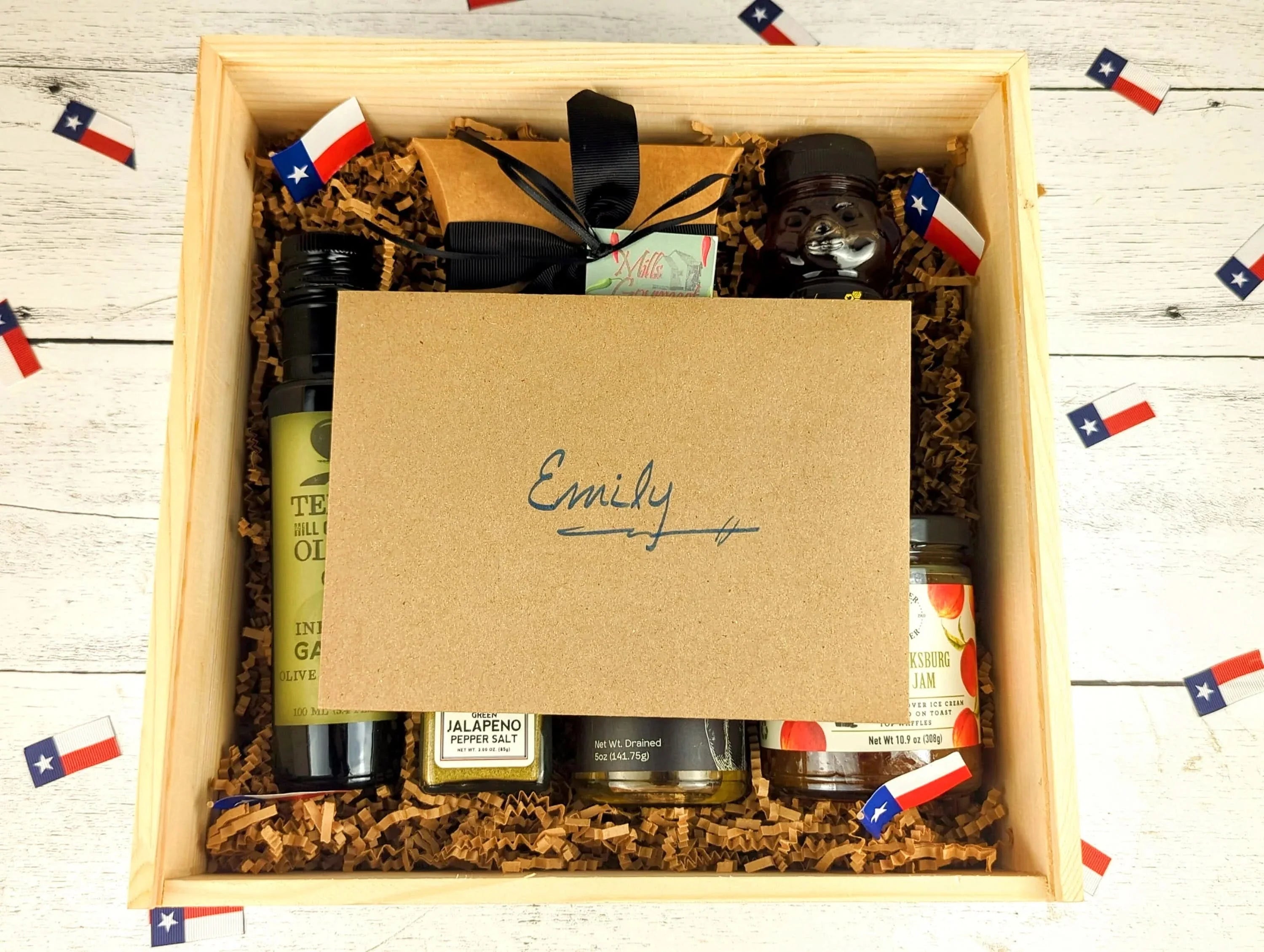 Open wooden gift box filled with Texas-made gourmet items, including infused olive oil, jam, seasoning, and sweets, topped with a personalized kraft card addressed to Emily, arranged with shredded paper filler and Texas flag confetti.
